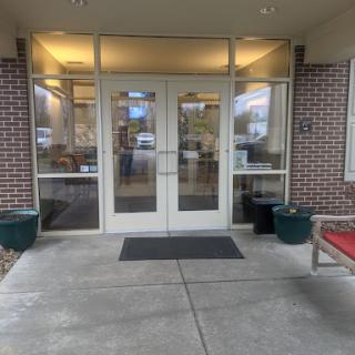 The entrance of a building featuring double doors and large windows, flanked by planters and a bench.