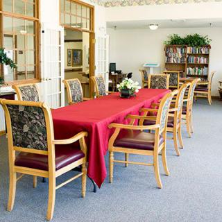 A well-lit dining area with a long table covered in a red tablecloth, surrounded by wooden chairs and a bookshelf in the background.