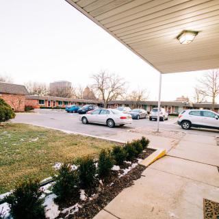 A parking lot with several cars parked near a building, featuring a covered walkway and trees in the background.