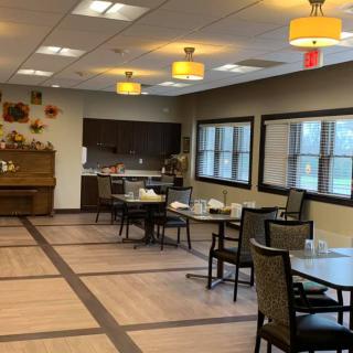 An empty dining room with neatly arranged tables and chairs, featuring a kitchen area and large windows.