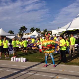 A lively outdoor event featuring volunteers in bright neon shirts and a person dressed in a colorful hamburger costume.