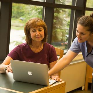 A woman is receiving guidance on using a laptop from another person in an indoor setting.
