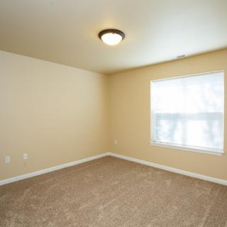 An empty room with beige walls, a window, carpeted floor, and a ceiling light fixture.