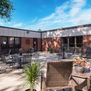 An outdoor patio area with tables, chairs, and plants against a brick building backdrop under a blue sky.