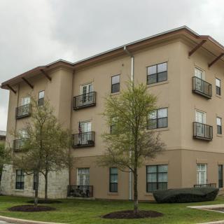 A modern three-story apartment building with balconies, surrounded by trees and grass.