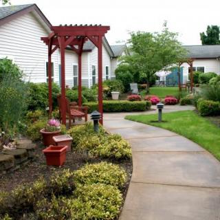 A beautifully landscaped garden with a winding pathway, red arbor, and benches surrounded by lush greenery and shrubs.