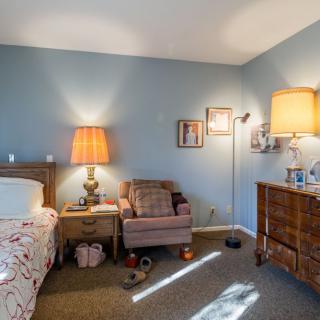 A cozy bedroom with a bed, nightstand, and comfortable chair illuminated by warm lights.