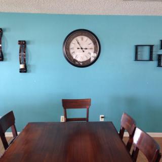 A dining room with a wooden table and chairs, featuring a blue wall adorned with a clock and decorative items.