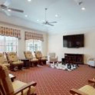 A spacious living room with a red carpet, lined with comfortable chairs, large windows, a ceiling fan, and a television mounted above a fireplace.