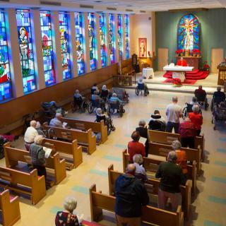 A church interior with congregants seated in pews, featuring stained glass windows and an altar at the front.