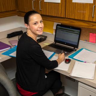 A woman is working at her desk, smiling while using her laptop.