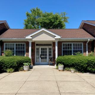 A brick residential building with a central entrance, flanked by large windows and surrounded by greenery.