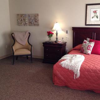 A cozy bedroom with a neatly made bed, a nightstand with a lamp, and an elegant chair.