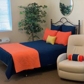 A cozy bedroom with a neatly made bed, colorful pillows, a cushioned chair, and a potted plant.