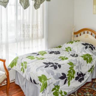 A cozy bedroom featuring a single bed with floral bedding, a chair, and large window with curtains.