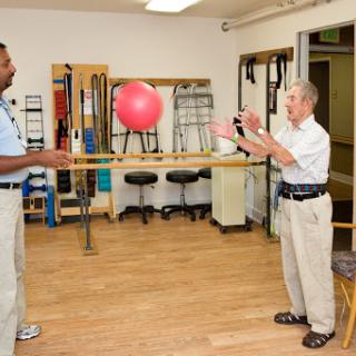 A physical therapy session where an elderly person practices catching a ball with the assistance of a therapist.