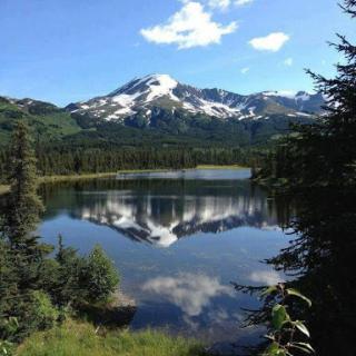 A serene view of a mountain reflected in a tranquil lake, surrounded by lush greenery and a clear blue sky.