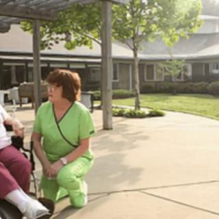 A nurse kneels beside an elderly person in a wheelchair, engaging in conversation outdoors.