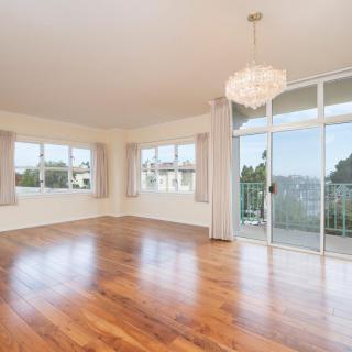 A spacious, empty room with hardwood flooring, large windows, and a chandelier, offering plenty of natural light and access to a balcony.