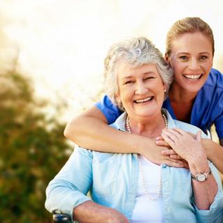 A happy elderly woman with a younger woman embracing her from behind outdoors.