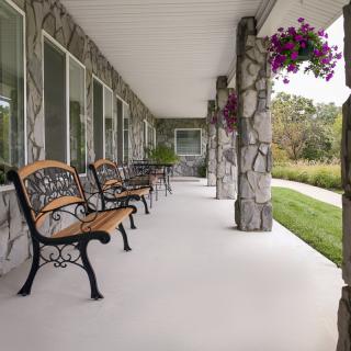 A porch with stone columns and benches, adorned with hanging flowers and surrounded by greenery.