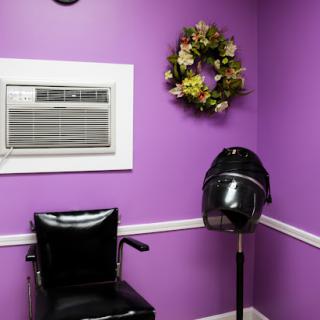 A cozy hair salon corner featuring a black chair, a vintage hair dryer stand, and vibrant purple walls adorned with a floral wreath.