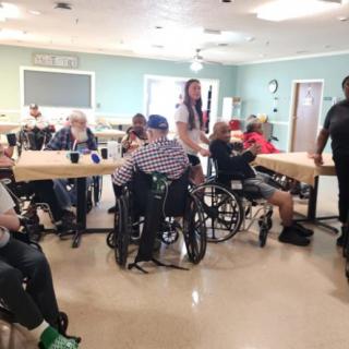 A group of elderly individuals and caregivers are gathered in a community room of a nursing home, engaging in conversation and activities.