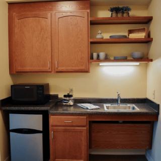 A small kitchen area with wooden cabinets, open shelves, a microwave, mini fridge, and sink.