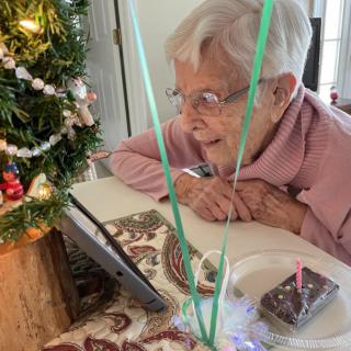 An elderly woman smiling during a video call, celebrating with a small birthday cake and Christmas decorations.