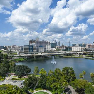 A vibrant cityscape with a lake and park in the foreground under a sky filled with clouds.