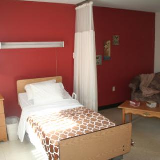 A simple hospital room featuring a single bed, a chair, and red walls.