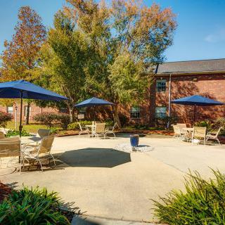 A sunny patio area with tables, chairs, and large umbrellas surrounded by trees and a building.
