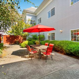 A sunny patio area with a red umbrella and matching outdoor furniture, surrounded by greenery.
