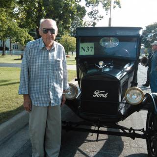 An elderly man stands beside a vintage Ford car on a sunny day.