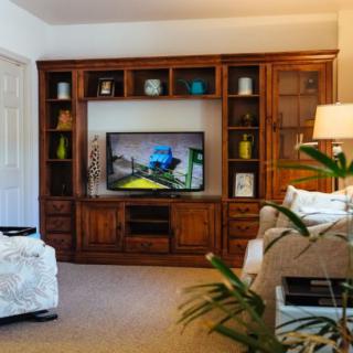 A cozy living room with a wooden entertainment center and a television surrounded by decorative shelves, lamps, and plants.