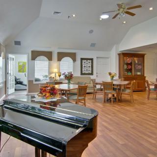 A spacious living and dining area featuring a grand piano, wooden flooring, and ceiling fans, with ample natural light from large windows.