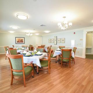 A well-lit dining room with neatly arranged tables and chairs, featuring chandeliers and wooden flooring.