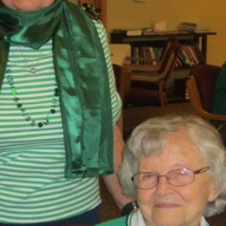 An elderly woman with glasses is smiling indoors, next to a person wearing a green scarf and striped shirt.