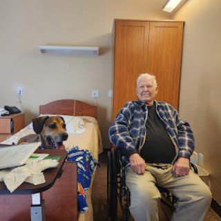 An elderly man in a wheelchair smiles alongside a dog in a care facility room.