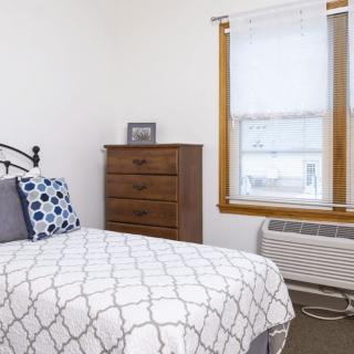 A cozy bedroom with a neatly made bed, a dresser, and a window with an air conditioning unit.