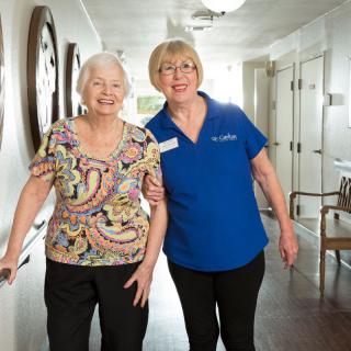 An elderly woman walking with assistance from a caregiver in a nursing home hallway.