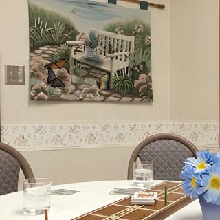 A dining area with a decorated table, featuring a floral centerpiece and wall art depicting a garden scene.