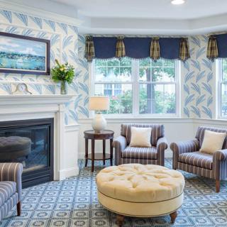 A cozy living room with a fireplace, featuring striped chairs, an ottoman, and floral wallpaper.