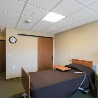 A hospital room with a single bed and an overbed table, featuring a clock on the wall and a wooden sliding door.