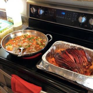A stove with a foil pan of cooked meat and a skillet of vegetables.