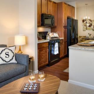 A cozy apartment interior featuring a living room with a sofa and a lamp, adjacent to a kitchen with wooden cabinets.