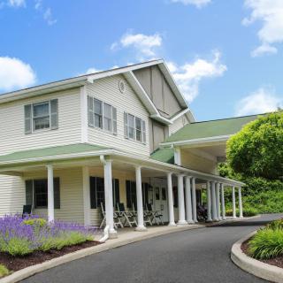 A two-story residential building with a green roof and a covered porch, surrounded by landscaped gardens on a sunny day.