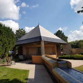 A modern pavilion with a distinctive pyramid roof surrounded by greenery and a flowing water feature.