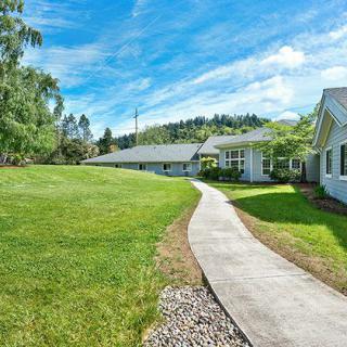 A scenic view of a pathway leading through a grassy area with buildings on either side and trees in the background under a clear blue sky.