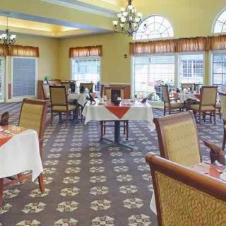 A spacious dining room with elegantly set tables and comfortable chairs, featuring large windows and chandeliers.